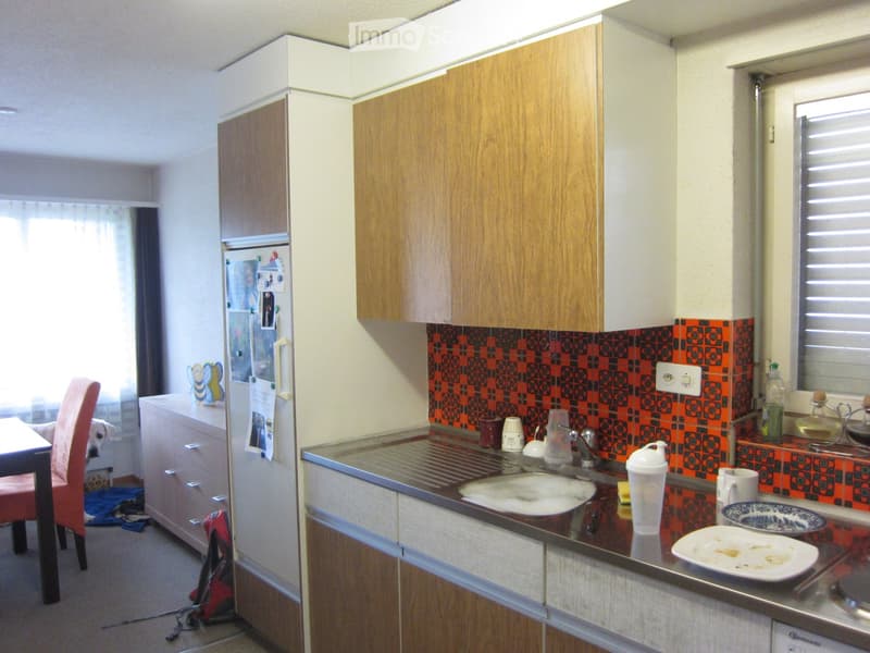 A small kitchen with wooden cabinets, a stainless steel sink, and various items on the counter including cups, a bottle, and a plate. There is a chair and table visible in the adjacent area.