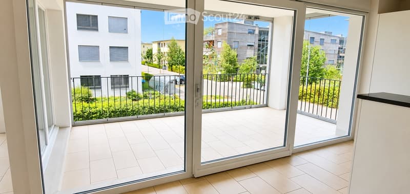 Bright, modern balcony with white tiled flooring, large glass sliding doors, metal railing, and a view of greenery and buildings.