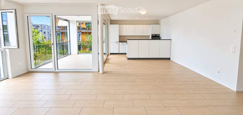 Bright, open-plan living and dining area with light wooden flooring, white kitchen cabinets, and a balcony with a metal railing.