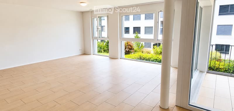 Empty room with light wooden flooring, white walls, and large windows offering a view of greenery outside.