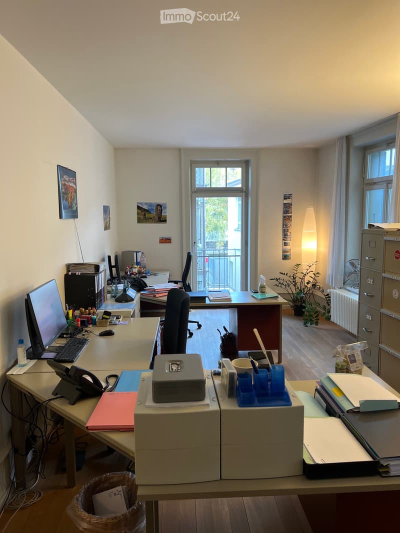 Office space with desks, chairs, computers, and office supplies. Features large windows, a plant, and filing cabinets.
