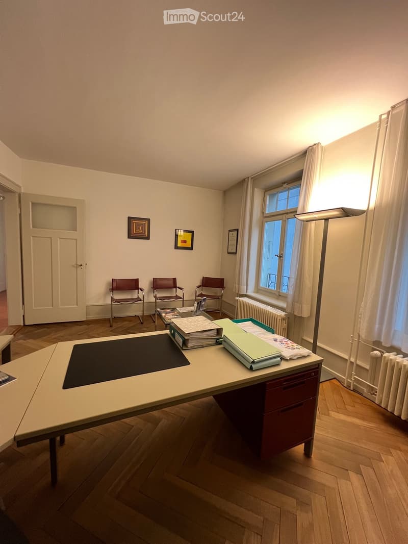 Office space with wooden flooring, large window with white curtains, a table with a black mat, three chairs, a radiator, a lamp, and framed pictures on the wall.