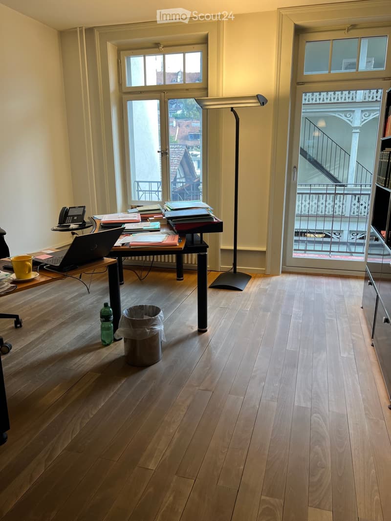 Office space with wooden flooring, large windows, and a balcony view. Includes a desk with a laptop, telephone, and various office supplies, along with a floor lamp and a trash can.