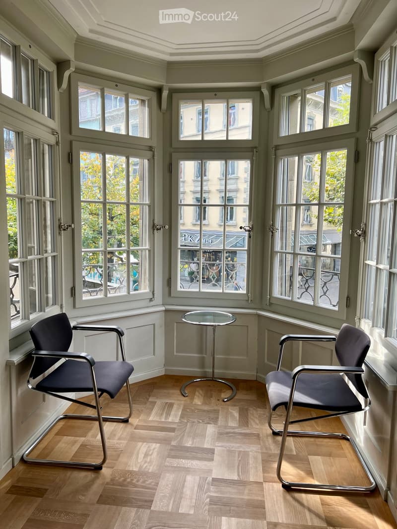 Brightly lit room with large windows, wooden floor, two chairs, and a small round table.
