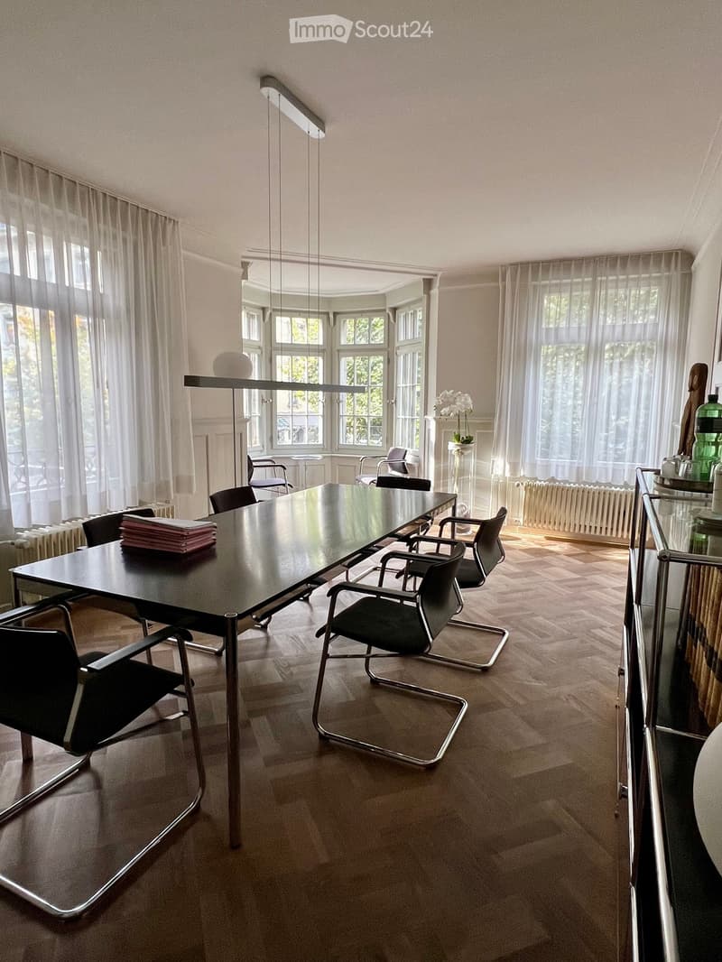 Modern office or meeting room with a large black conference table, multiple black chairs, large windows with sheer curtains, and a hanging light fixture.
