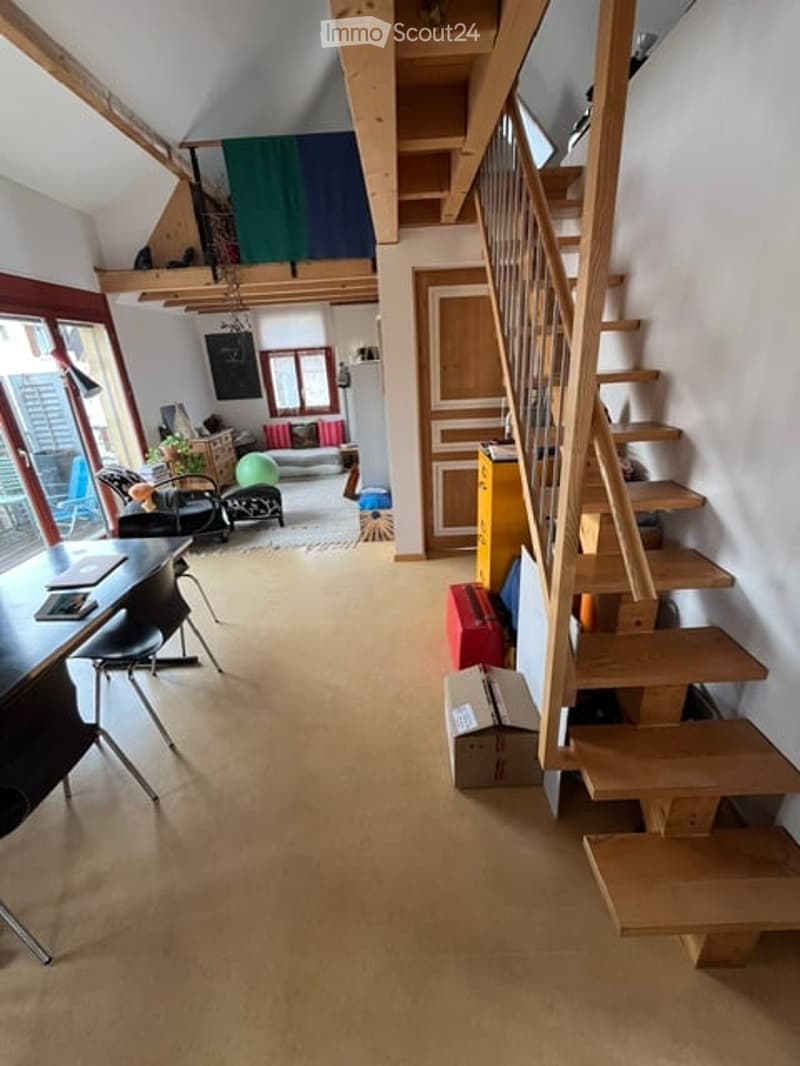 The image shows a modern, open-plan living and dining area with exposed wooden beams on the ceiling. The space features a wooden staircase leading to an upper level, a dining table with chairs, a sofa, a small ball, and a lamp. The room has large windows