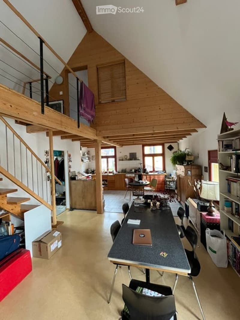 Open-plan living and dining area with exposed wooden beams, a staircase leading to an upper level, and a kitchen area visible in the background. The space includes a dining table with chairs, a lamp, and bookshelves.
