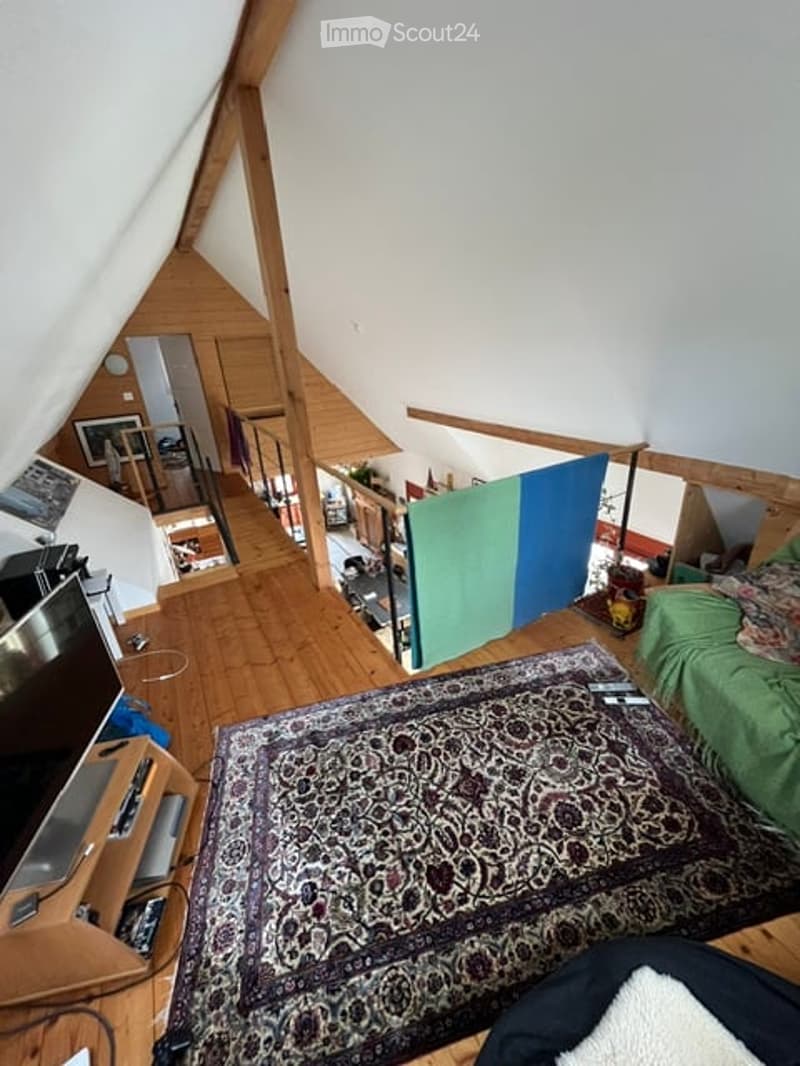 Attic room with wooden floors, slanted ceilings, and exposed wooden beams. Features a patterned rug, a green bed, a TV stand, and a wooden shelf.