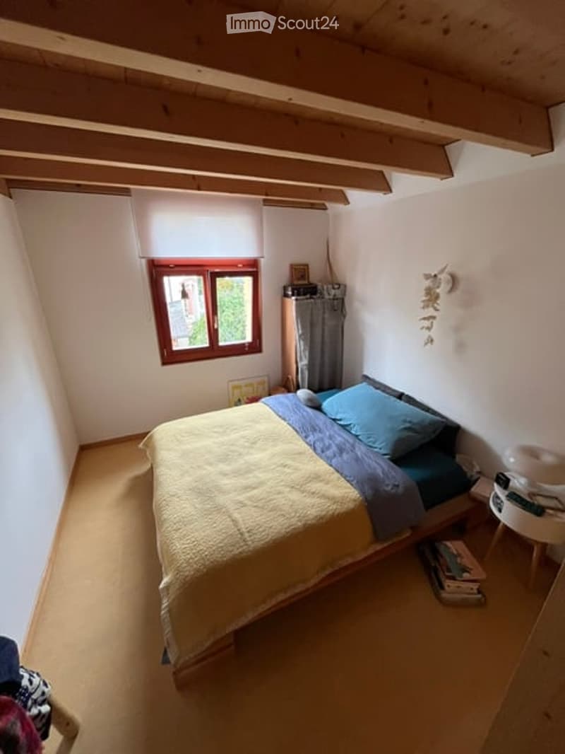 Cozy bedroom with wooden ceiling beams, a single bed with blue and yellow bedding, a small nightstand, and a window with a curtain.