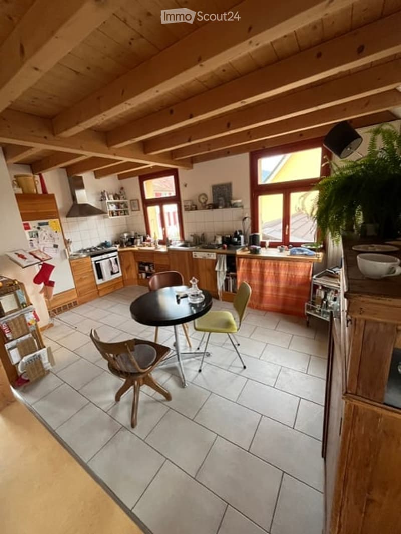 Bright kitchen with wooden beams, white tiled floor, and wooden cabinets. Features a central dining table with chairs, a stove, oven, dishwasher, and a potted plant on the counter.