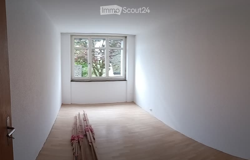 Empty room with white walls, wooden floor, and a single window showing greenery outside. No furniture or equipment present.