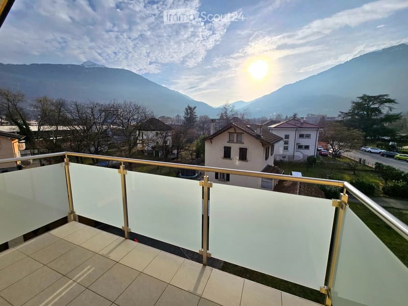 Balcony with glass railing, tiled floor, overlooking a town with houses and mountains in the background.