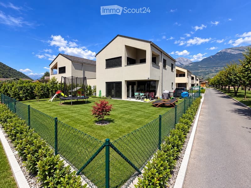 A modern residential complex with a well-maintained lawn, a trampoline, a slide, outdoor furniture, and lounge chairs, surrounded by a green fence and trees, with mountains in the background.