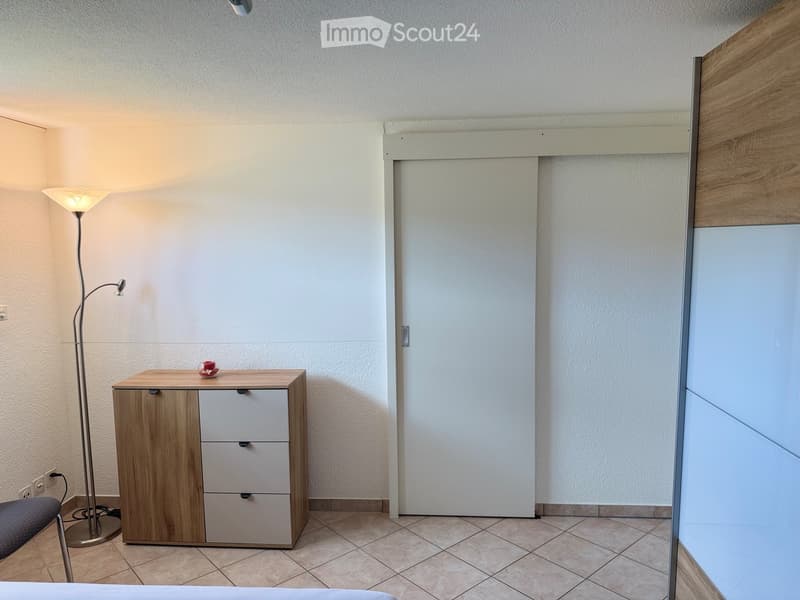 A small, furnished room with a wooden cabinet, a floor lamp, and a chair. The room has tiled flooring and white walls.