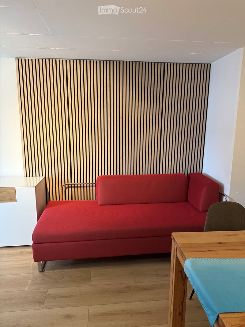 A modern living room with a red sofa, a wooden table with a blue cloth, and a chair. The room features a wooden floor and a striped wall panel.