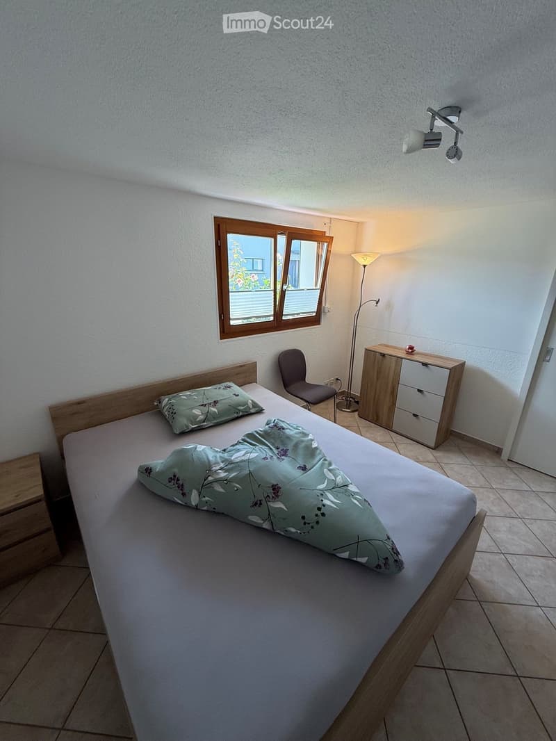 Cozy bedroom with a wooden bed, green floral bedding, a wooden nightstand, a chair, and a floor lamp near a window with wooden frames.