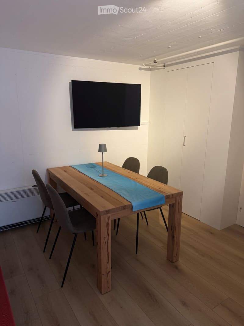 A modern, minimalist room with a wooden dining table, four chairs, a small lamp, and a mounted screen on the wall.