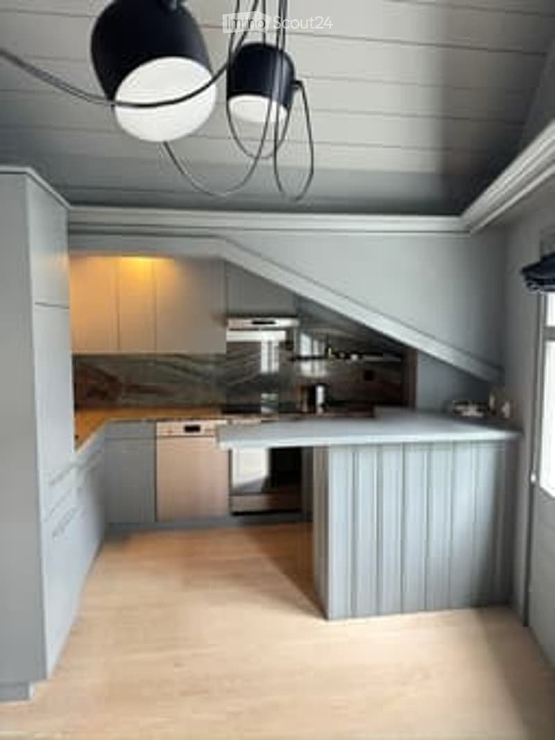 Modern kitchen with light grey cabinetry, a central island, and stainless steel appliances including a stove and dishwasher. The space features a sleek, minimalist design with a hanging pendant light fixture above the island.