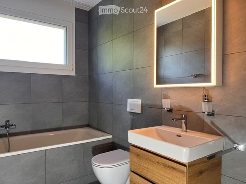 Modern bathroom with dark grey tiles, a bathtub, toilet, sink, and illuminated mirror.