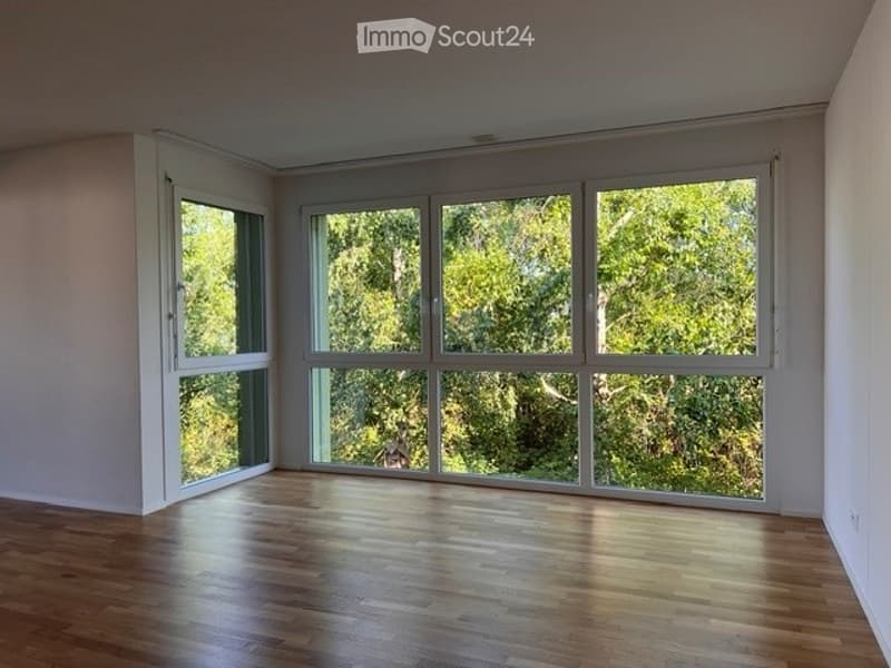 Empty room with wooden flooring and large windows offering a view of greenery outside.