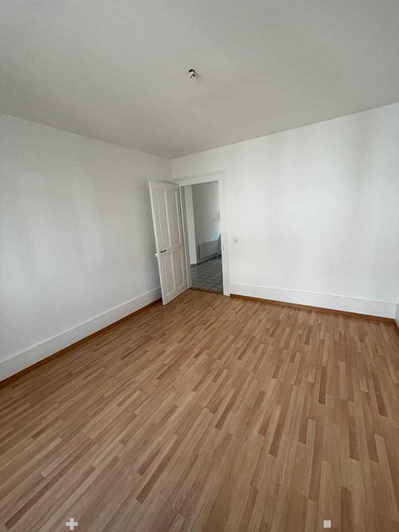 Empty room with light-colored walls and wooden flooring, featuring an open doorway leading to another room.