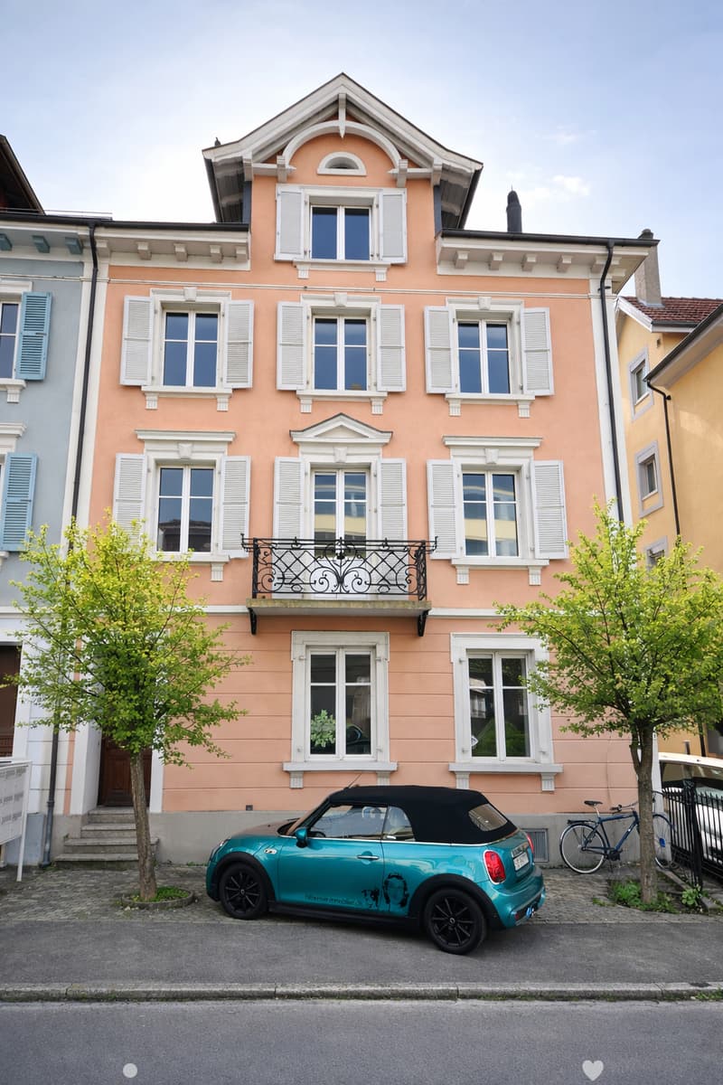 A two-story pink building with white window frames and shutters, featuring a balcony with a decorative railing. A turquoise convertible car is parked in front, and a bicycle is leaning against a fence.