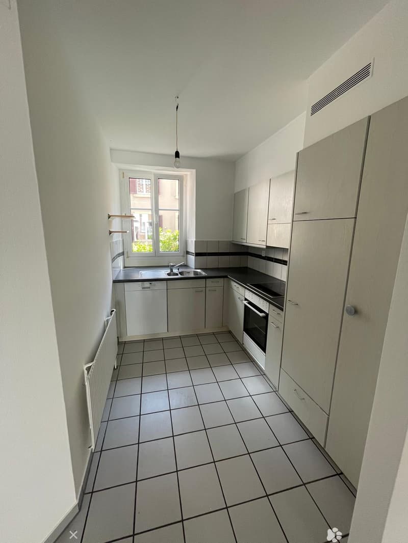 Modern kitchen with white cabinets, tiled floor, sink, oven, and dishwasher. Natural light from a window on the left.