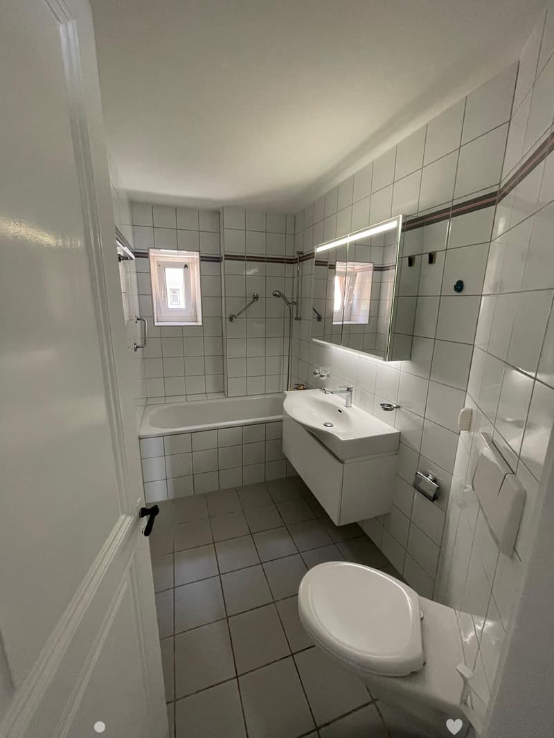 Modern bathroom with tiled walls and floor, featuring a bathtub, sink, toilet, and large mirror with built-in lighting.