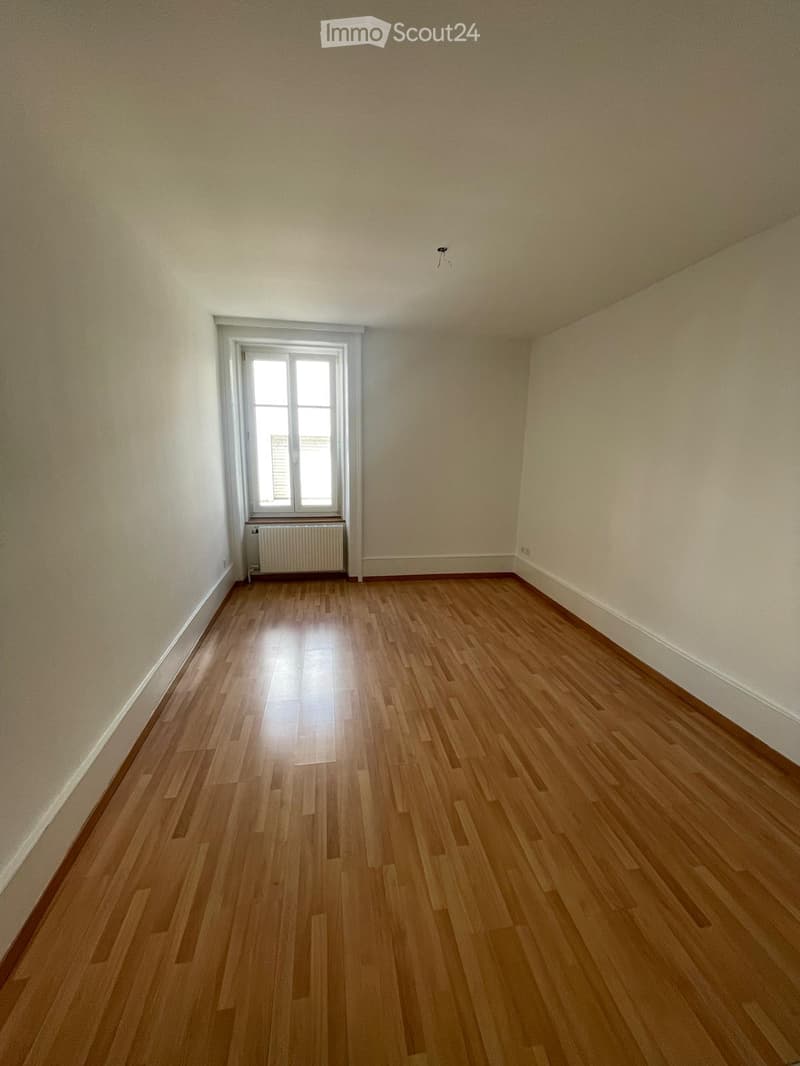 Empty room with wooden flooring, white walls, and a single window with a radiator below it.
