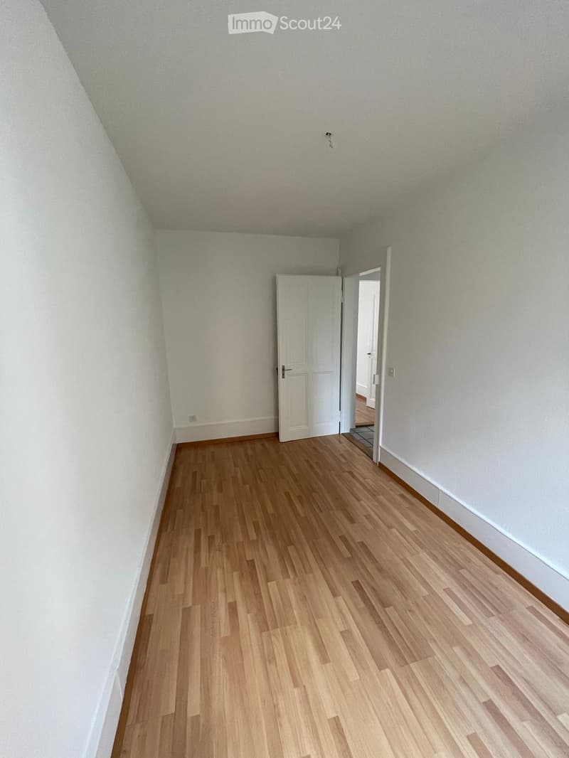 Empty room with light wooden flooring and white walls, featuring a white door leading to another space.