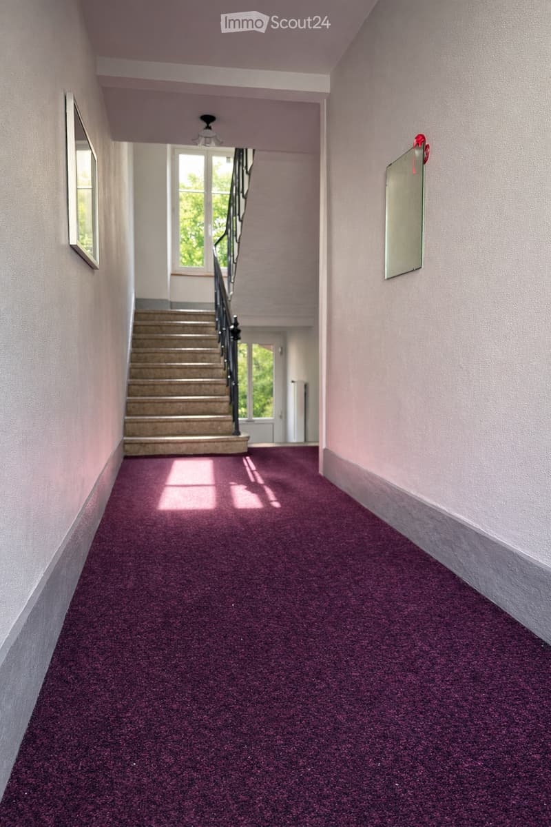 A carpeted hallway with light pink walls, a staircase leading upwards, and a window at the end allowing natural light to enter. The carpet is a deep purple color, and the hallway features a simple ceiling light fixture.