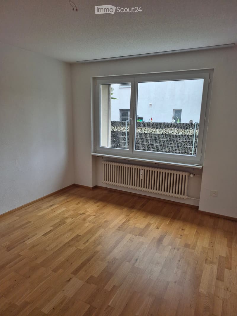 Empty room with wooden flooring, white walls, a radiator, and a large window with a view of a stone wall and outdoor area.