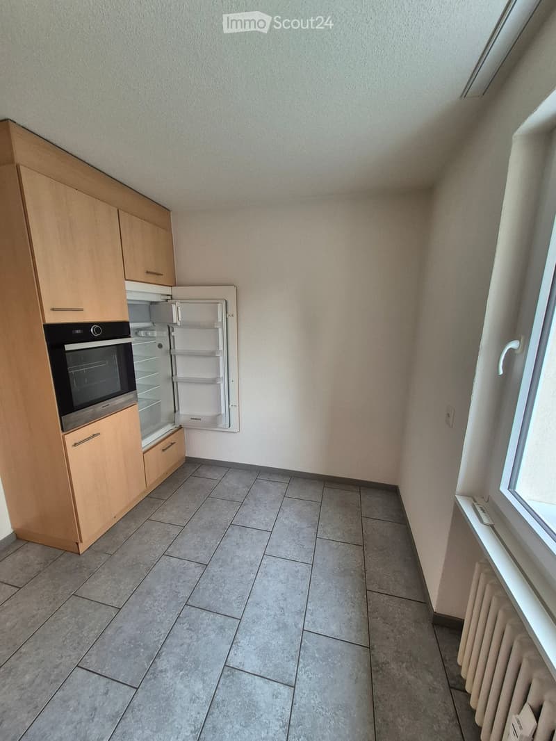 Modern kitchen with light wooden cabinets, stainless steel oven, and refrigerator. Grey tiled floor and white walls.