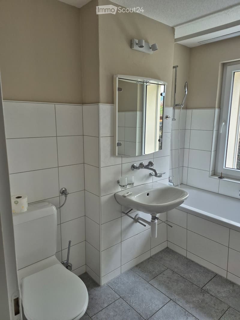Modern bathroom with white tiles, a toilet, a sink, a bathtub, a mirror, and a window providing natural light.