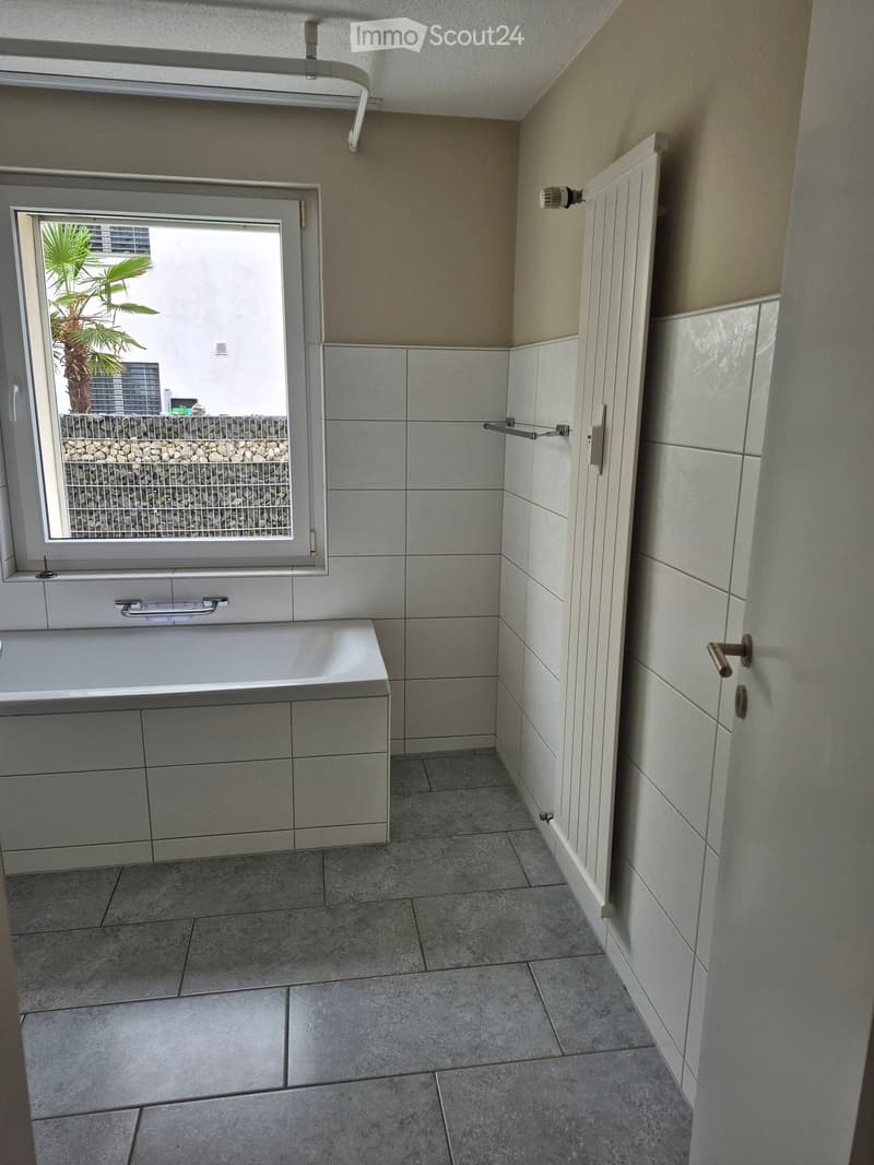 Modern bathroom with a bathtub, built-in cabinets, tiled walls, and a window overlooking a small outdoor area with a palm tree.