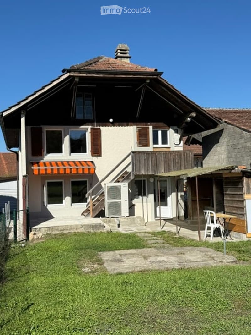 Two-story house with a wooden balcony, red-tiled roof, and a small outdoor seating area with a table and chairs.