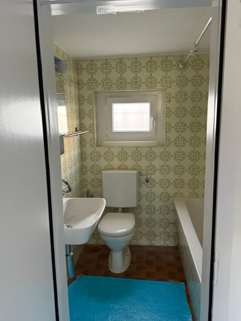 A small bathroom with a toilet, sink, and bathtub. The walls are covered in a green patterned wallpaper, and there is a small window above the toilet. A blue bath mat is placed on the floor in front of the bathtub.