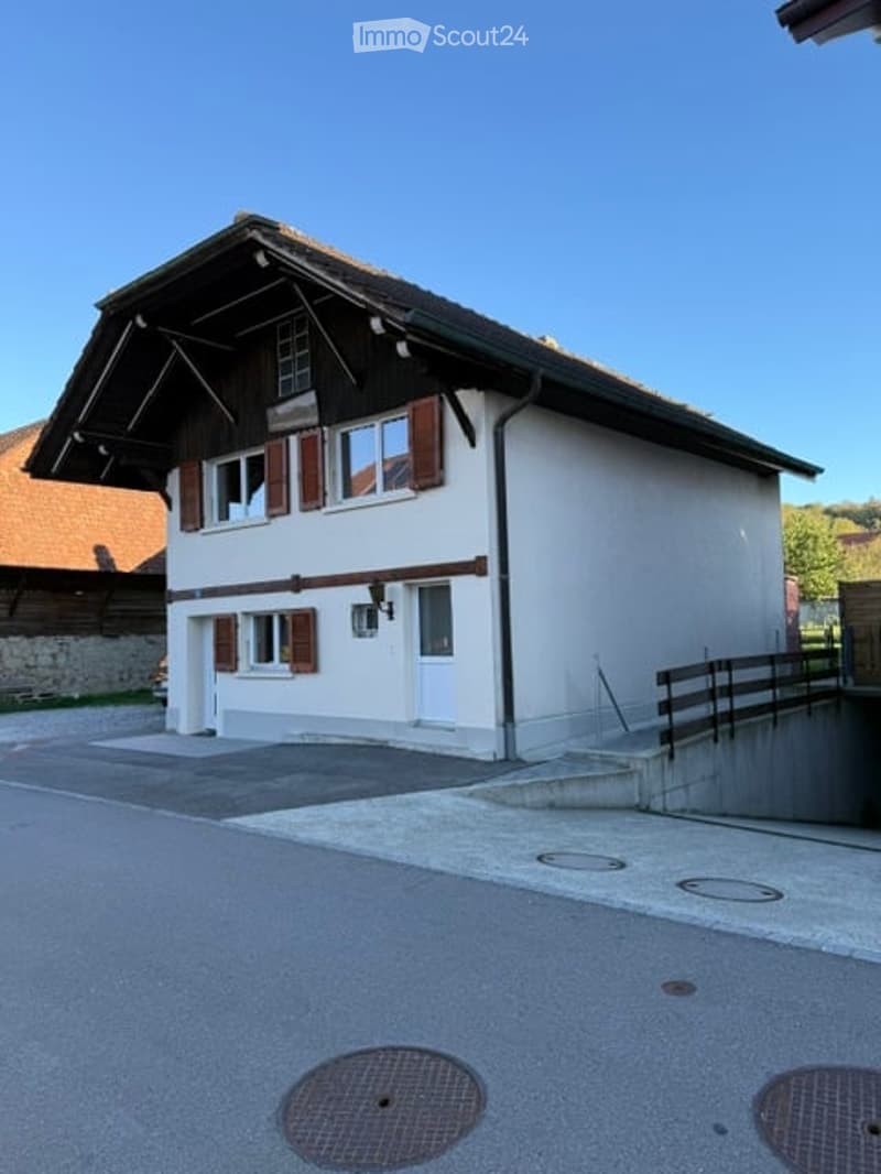 A single-story house with white walls, brown shutters, and a wooden roof. The house features multiple windows and a small porch area with a wooden railing.