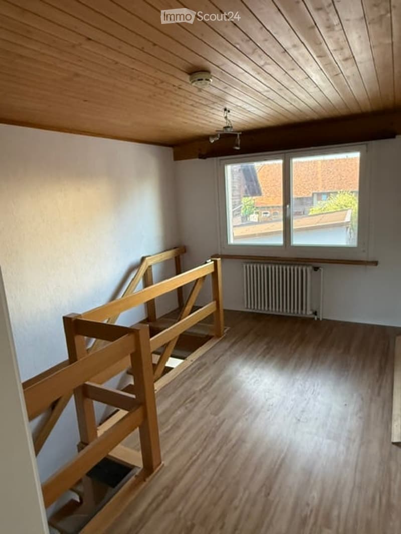 Empty room with wooden flooring, white walls, and a window with a view of neighboring houses. A wooden staircase railing is visible, and a radiator is placed under the window.