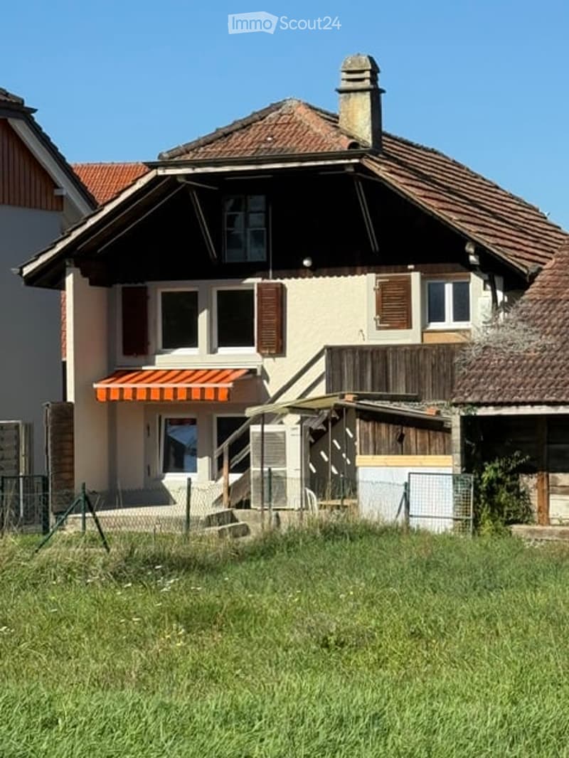 Two-story house with a tiled roof, beige walls, and brown shutters. Features include a small awning over the entrance and a staircase leading to a raised ground floor.