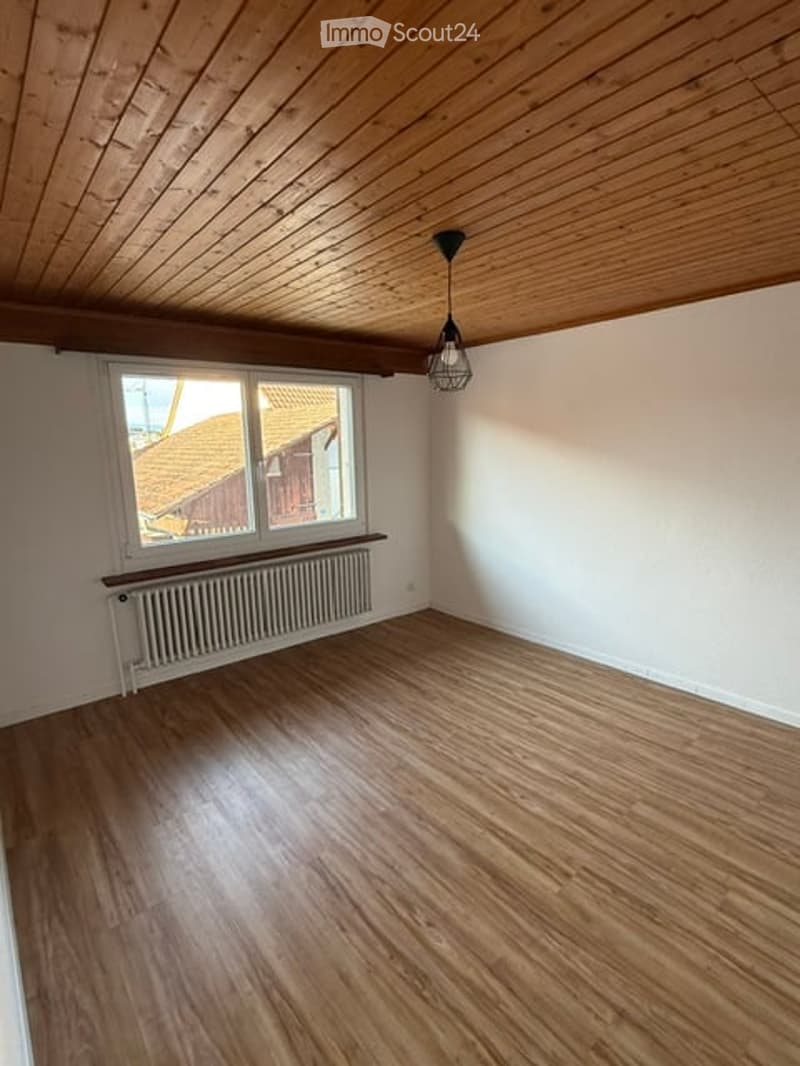 Empty room with wooden flooring, white walls, a window with a view of a roof, a radiator, and a hanging light fixture.
