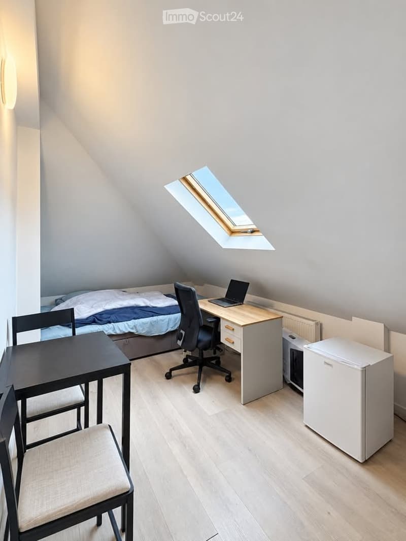 Cozy attic bedroom with a bed, desk, laptop, chair, small table, and mini-fridge. Features a skylight window and light wooden flooring.