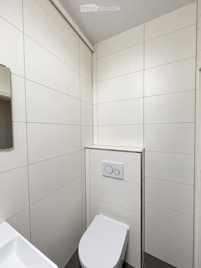 Modern bathroom with white tiled walls, a toilet, a sink, and a mirror. The space is clean and contemporary with a sleek design.