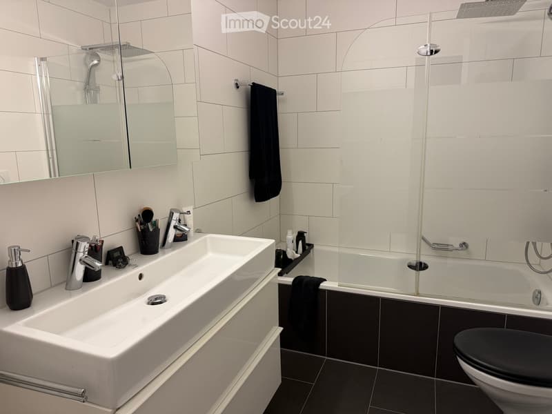 Modern bathroom with a double sink, bathtub, toilet, and shower area. Features include a large mirror, towel rack, and various toiletries.