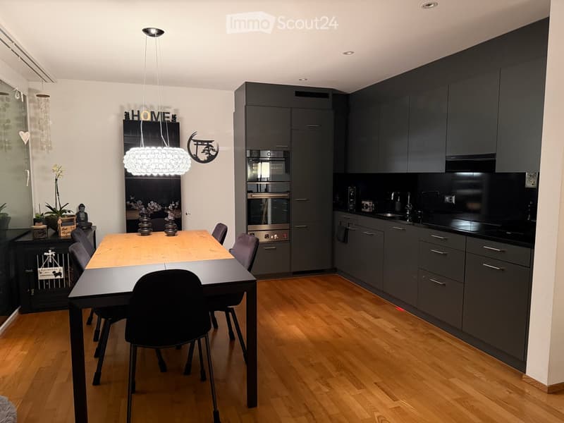 Modern kitchen and dining area with dark grey cabinets, a wooden dining table, and black chairs. Features a built-in oven, sink, and decorative items including vases and a light fixture.