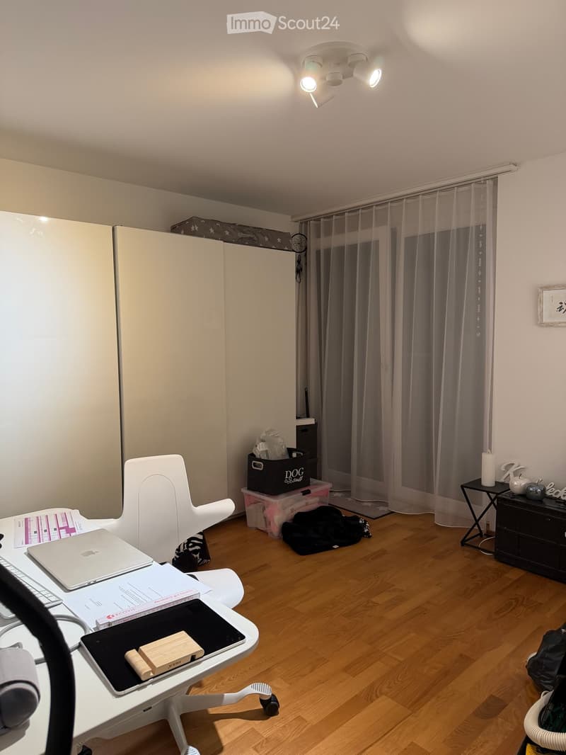 A modern, well-lit room with wooden flooring, a white desk with a laptop, a white chair, a small black table with a candle, and a window with sheer curtains.