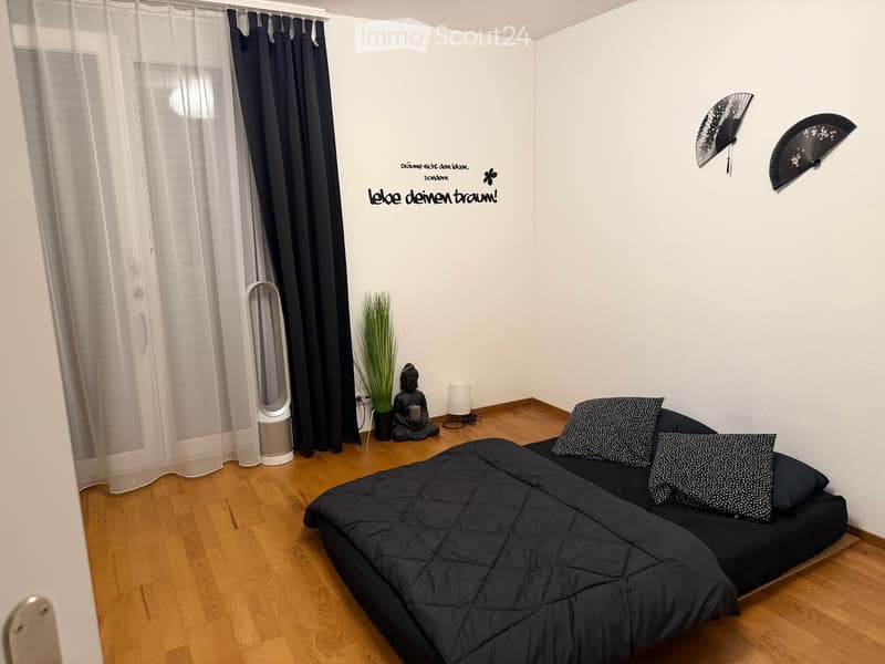 A modern, minimalist bedroom with a black sofa, decorative pillows, a small plant, a Buddha statue, and a wall-mounted fan. The room features wooden flooring and large windows with sheer curtains.