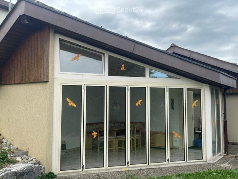 Modern single-story house with large glass sliding doors leading to a patio area. The interior shows a dining area with a table and chairs visible through the glass.
