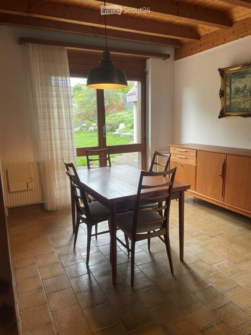 Bright dining room with wooden dining table and chairs, large window with curtain, wooden cabinet, and a framed picture on the wall.