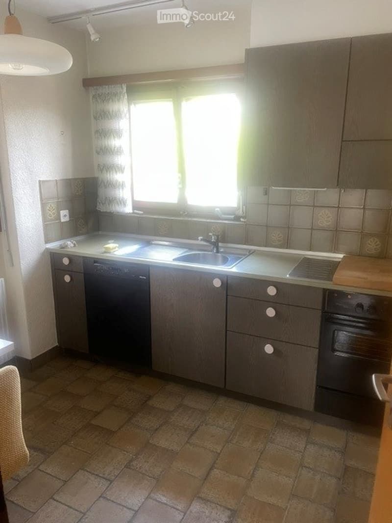 Kitchen with dark wooden cabinets, tiled floor, sink, dishwasher, and oven. Natural light from a window with curtains.